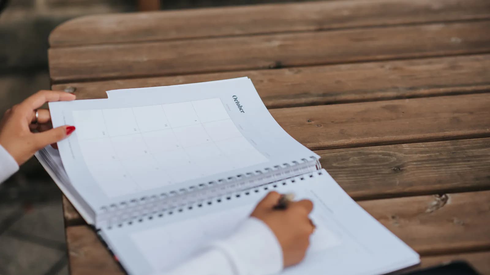 An open monthly desk planner next to a notebook, a pen and a cup of coffee on a workspace.