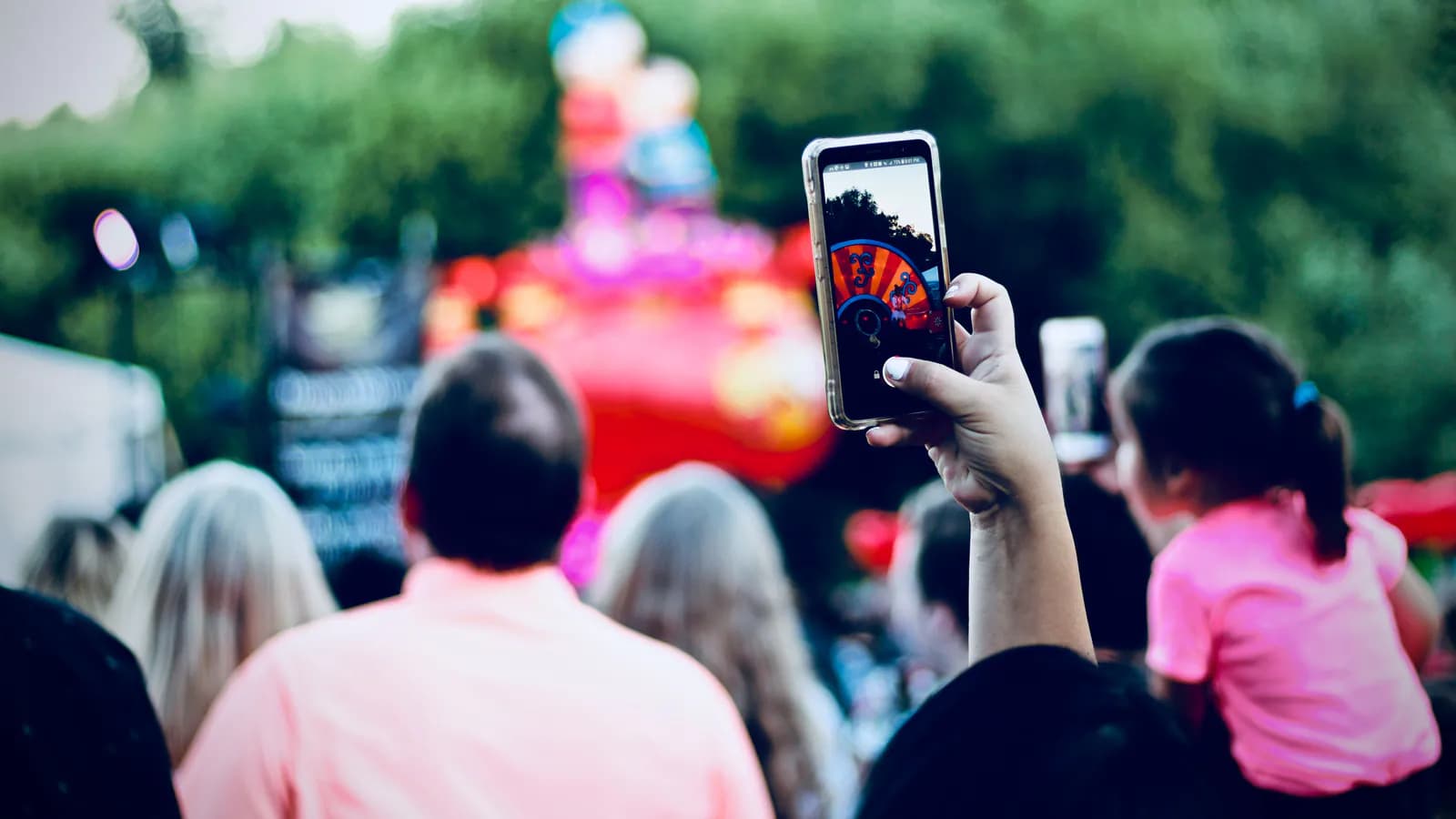 At an outdoor event, a raised hand in the crowd films a bright, colourful attraction on a phone.