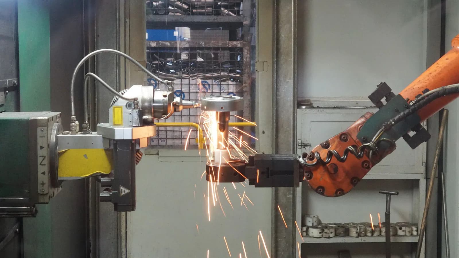 Sənaye robot qolu emalatxanada metal hissəni emal edir, ətrafa qığılcım səpələnir — avtomatlaşdırılmış proses insan nəzarəti altında işləyir.