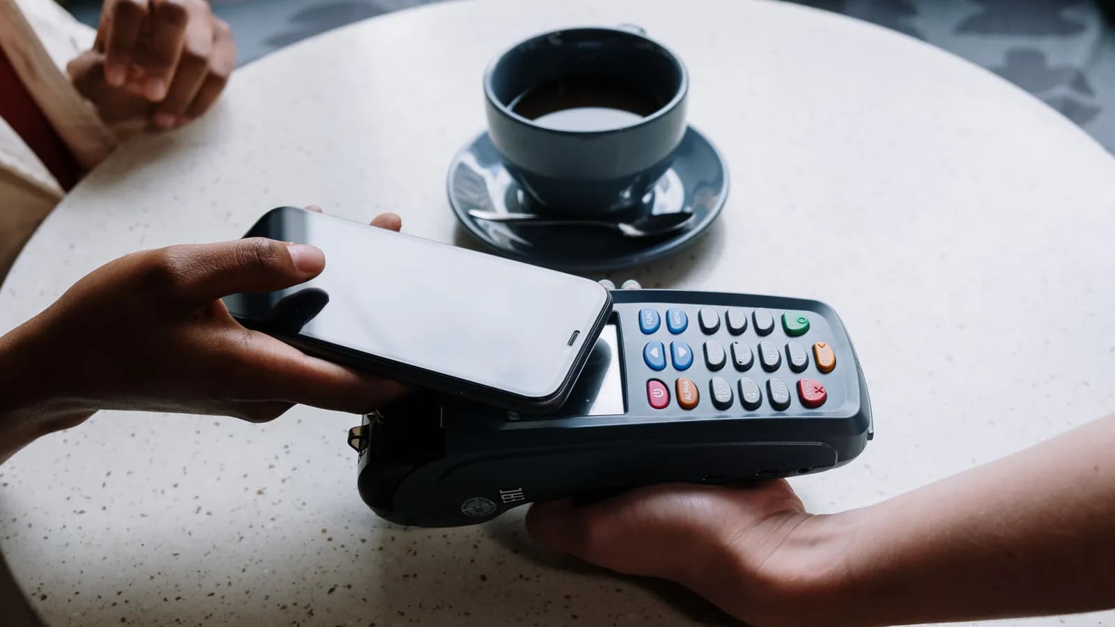 Kafedə bir müştərinin telefonunu ödəniş terminalına toxundurduğu, kassirin terminalı əlində tutduğu an — kontaktsız ödənişin arxa fonunda bir fincan qəhvə görünür.