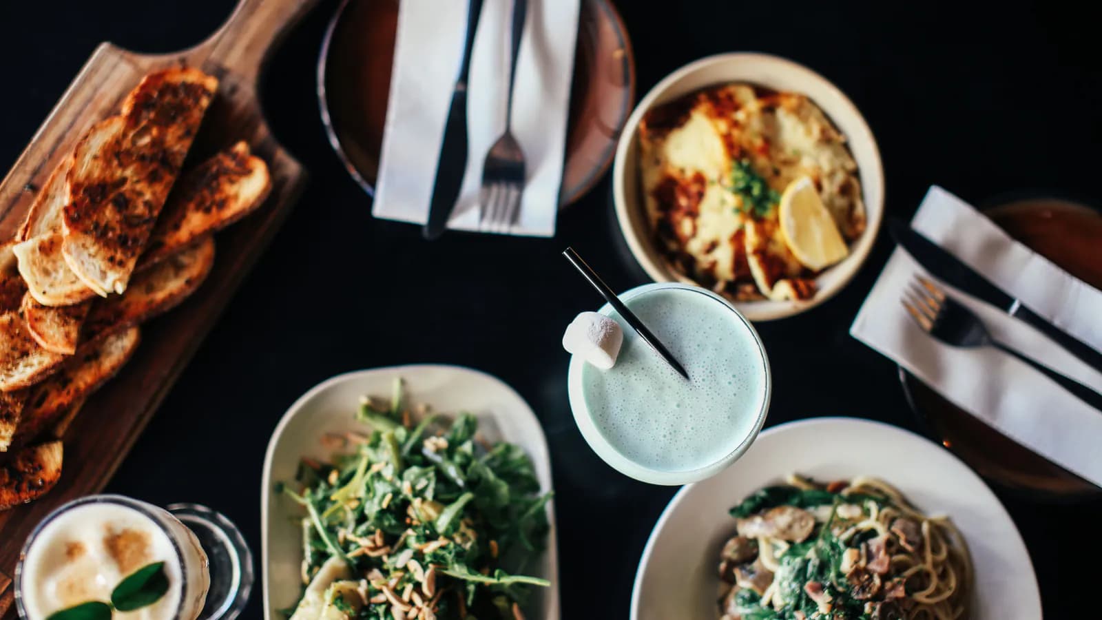 Restoran masasının yuxarıdan çəkilmiş görüntüsü, üzərində bir neçə fərqli yemək boşqabı və içki stəkanları düzülüb.