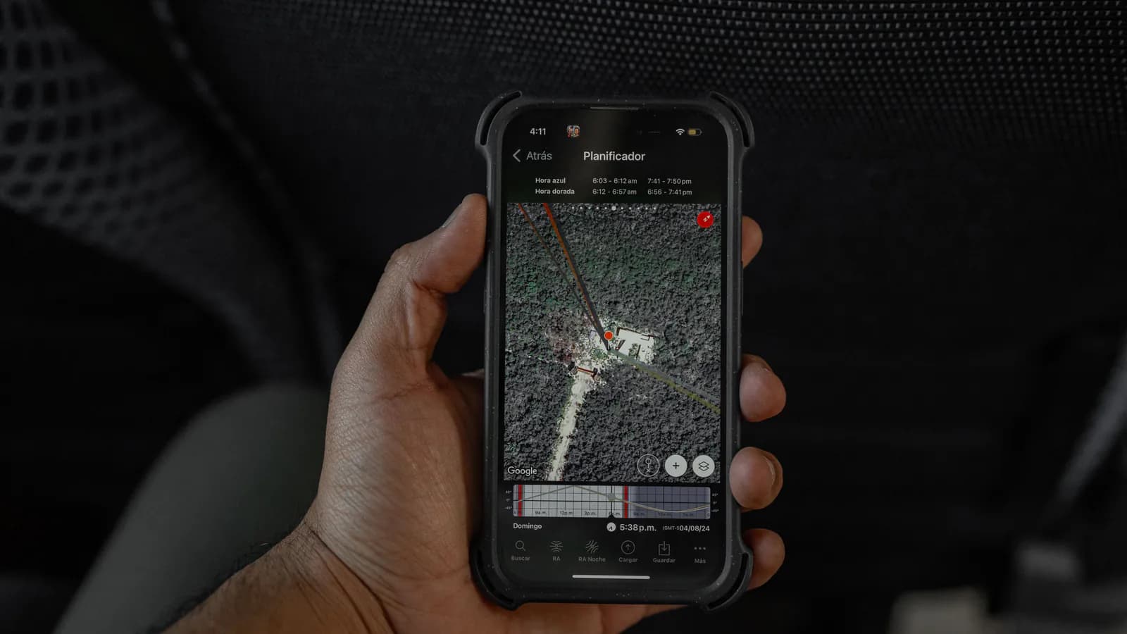 A hand holds a smartphone showing a satellite map with a red location pin marked on it, with a planning chart along the bottom of the screen.